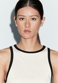 Sleeveless top in cream with a black ribbed neckline. Smooth fabric texture, fitted design, showcasing a clean and minimalistic aesthetic.