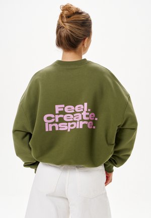 Kobieta z włosami związanymi w kok, ubrana w oliwkowozieloną bluzę z różowym napisem „Feel. Create. Inspire.” na plecach, w połączeniu z białymi spodniami.