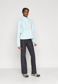 Αθλητικό ανοιχτό γαλάζιο τοπ με φερμουάρ και ψηλό γιακά, με αντιθετικές ραφές, σε συνδυασμό με σκούρο γκρι παντελόνι και πολύχρωμα αθλητικά παπούτσια.