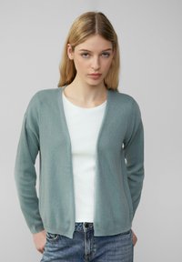 Hellgrüner Cardigan aus strukturiertem Stoff, mit V-Ausschnitt, offener Front und langen Ärmeln, getragen über einem weißen Top und blauen Jeans.