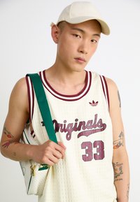 Bela mrežasta košarkaška dres s maroon akcentima, sa natpisom "Originals" i brojem "33." Zeleni remen na maloj beloj torbi.