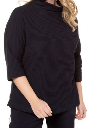 Kobieta w prążkowanym, ciemnogranatowym topie z rękawami do łokci i luźnym kroju, w połączeniu z ciemnymi spodniami z białym paskiem.