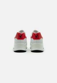 Vita sneakers med en röd glansig hälldetalj och svart logotyp. Rundad form, texturerad sula och vadderad krage. Baktill sett.