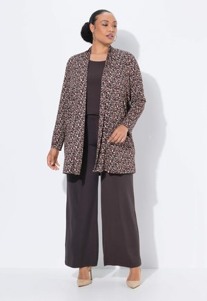 Vrouw in een open cardigan met luipaardprint, zwarte wijde broek en beige hoge hakken, staand tegen een witte achtergrond.