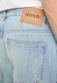 Šviesiai mėlynos džinsinės kelnės turi rudą odinį ženklelį su įspaustu „HUGO“. Siuvinėjimas matomas palei siūles ir kišenes, rodantis tvirtą dizainą.