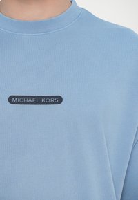 Világoskék pulóver, kerek nyakkivágással. Fekete téglalap alakú logó található rajta, a "MICHAEL KORS" fehér szöveggel középen a mellkasán.
