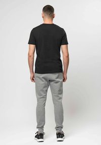 Mand stående med ryggen mod, iført en sort t-shirt, grå joggingbukser og sorte sneakers mod en ensfarvet hvid baggrund.