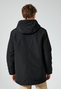 Parka negra con capucha y un acabado suave y mate. Presenta un corte relajado, mangas largas y una longitud hasta la mitad del muslo, adecuada para el uso casual.
