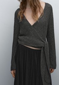 Femme portant un pull enveloppant gris foncé avec un lien sur le côté et une longue jupe plissée noire, se tenant devant un fond gris clair uni.