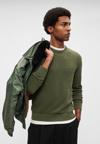 Alyvuogių žalios spalvos megztinis su tekstūruotu raštu, apvalia iškirpte ir ribotais rankogaliais, dėvimas po blizgia tamsiai žalia striuke.