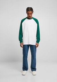 Langarm-Raglan-T-Shirt mit grünen Ärmeln und weißem Körper, kombiniert mit blauen Jeans und weißen Sneakers, das eine entspannte Passform zeigt.