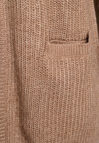 Maglione lavorato a maglia color marrone chiaro, con una texture a coste e una piccola tasca integrata sul lato inferiore. Aspetto morbido e accogliente.