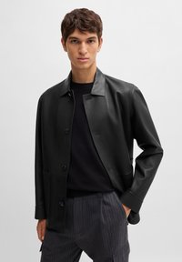 Black leather jacket with a collared design, front button closure, and side pockets. Worn over a dark sweater and pinstriped pants.