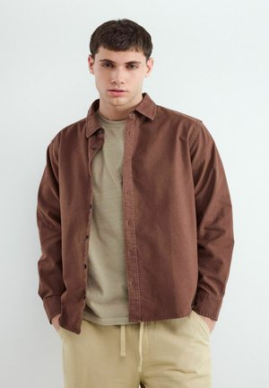 Mladý muž, ktorý má na sebe hnedú košeľu s gombíkmi cez béžové tričko a svetlo béžové nohavice, stojí s rukami v vreckách pri jednoduchej stene.