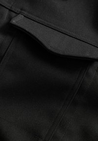 Material negru cu o suprafață texturată și o buzunare cu clapetă. Cusăturile vizibile și liniile clare creează un design structurat.