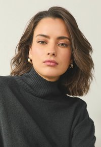 Melns augstas apkakles džemperis ar ribas tekstūru, izgatavots no mīksta materiāla. Modelim vidēja garuma viļņaini mati un apļveida auskari.