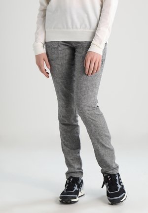 Osoba ubrana w dopasowane szare spodnie, czarno-białe trampki oraz biały długi rękaw, stojąca na jednolitym jasnym tle.