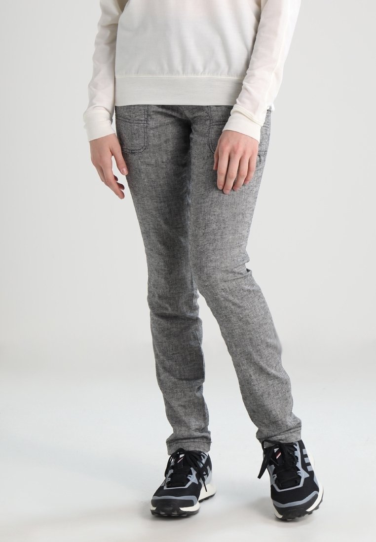 Personne portant un pantalon gris ajusté, des baskets noires et blanches, et un haut à manches longues blanc, se tenant sur un fond clair uni.