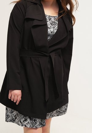 Femme portant un manteau noir ceinturé par-dessus une robe noire et blanche à motifs, avec des manches mi-longues et une coupe ample.