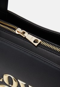 Musta strukturoidu käsilaukku teksturoidulla pinnalla, jossa on kultainen vetoketju ja etiketti, jossa on sõna "ARMAS" reljeefselt ofresseeritud.