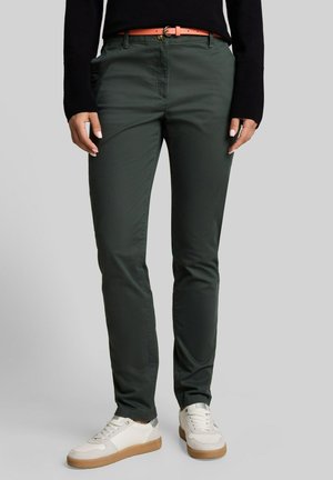 Personne portant un pantalon slim vert foncé avec poches avant, une ceinture corail, un haut noir à manches longues et des baskets blanches avec semelles beiges.
