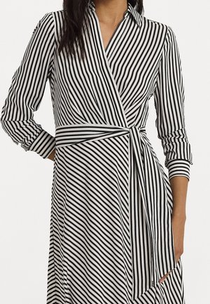 Femme portant une robe portefeuille rayée verticale noir et blanc avec des manches longues et une ceinture assortie nouée à la taille.