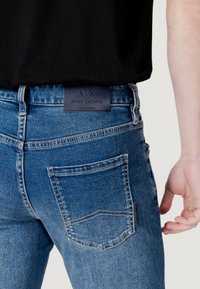 Jeans de mezclilla azul con un diseño ajustado, que presentan un bolsillo trasero visible y una etiqueta de Armani Exchange en azul oscuro en la cinturilla.