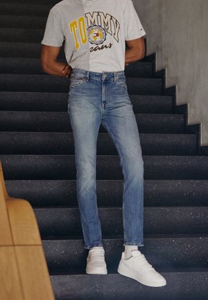 Osoba ubrana w jasnoniebieskie dżinsy, białe trampki i szarą koszulkę Tommy Jeans stojąca na ciemnych schodach z rękami za plecami.