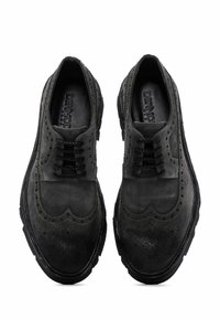 Chaussures en daim noir avec un design brogue, présentant des détails perforés, des lacets ronds et une semelle en caoutchouc texturée.
