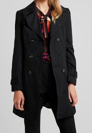 Femme portant un trench-coat noir croisé sur un chemisier coloré à motifs avec un nœud noué au col et un pantalon noir.