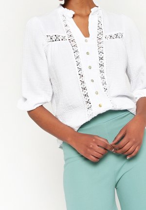 Kobieta w białej, teksturowanej bluzce z koronkową lamówką i guzikami, w połączeniu z jasnoturkusowymi spodniami z wysokim stanem, ręce splecione przy talii.