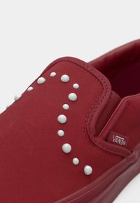 Røde lerrets slip-on sko med flat såle, med hvite nagledetaljer ordnet i et buet mønster på overdelen.