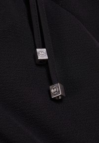 Černá tkanina s texturovaným povrchem má dva konce na stahovací šňůrce, z nichž každý má stříbrnou kovovou kostku s logem.