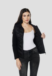 Črna puhasta jakna z vzorcem šivov, z zadrgo spredaj in kapuco. Oblečena čez belo majico brez rokavov in kombinirana s temnimi kavbojkami.