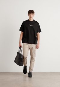 Zwarte katoenen t-shirt met logo, beige cargobroek met zijzakken, en zwarte sneakers. Draagt een zwarte totebag, wandelend over een neutrale vloer.