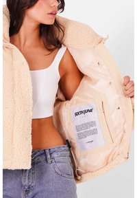 Beige faux-shearling jakke med en quiltet satinforing. Har trykknaplukninger og et mærke indeni. Båret over en hvid cropped top og jeans.