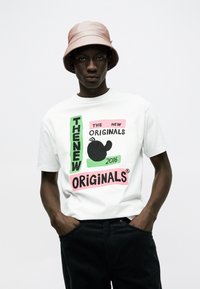 Mann trägt hellrosa Fischerhut, weißes T-Shirt mit buntem Schriftzug "THE NEW ORIGINALS 2016" und schwarze Hose, Hände in den Taschen.