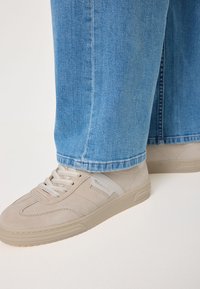 Sneakers beige in suede con lacci bianchi indossati con jeans in denim blu, mostrati di lato su una superficie chiara.