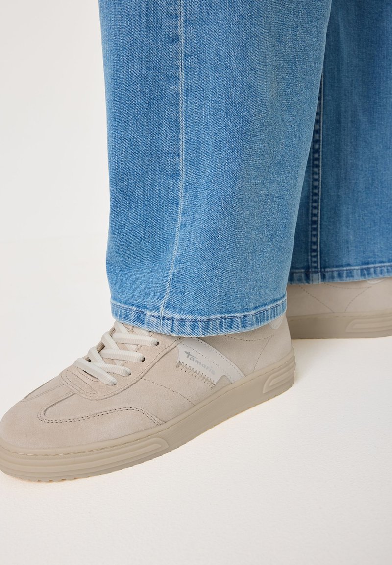 Sneakers beige in suede con lacci bianchi indossati con jeans in denim blu, mostrati di lato su una superficie chiara.