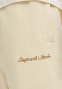 Lys beige joggers lavet af blødt stof, med et tonalt broderet logo "Originals Studio" i kursiv på låret.