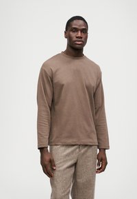 Muškarac u smešanoj košulji dugih rukava smeđe boje i bež teksturiranim hlačama stoji nasuprot jednostavnoj beloj pozadini.