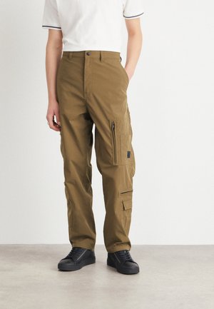 Pantaloni cargo color khaki con più tasche, taglio dritto, tessuto resistente, chiusure a zip sulle tasche laterali, indossati con sneakers nere.
