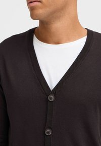 Cardigan marron foncé avec un col en V, présentant une texture côtelée et une fermeture à trois boutons à l'avant, superposé sur une chemise blanche.
