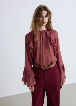 Donna con capelli ondulati che indossa una camicetta trasparente a maniche lunghe con volant e pantaloni a vita alta con cintura intrecciata su sfondo semplice.