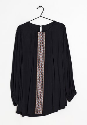 Czarna bluzka z długim rękawem z plisowanego materiału z kolorową geometryczną pionową lamówką z przodu, zawieszona na czarnym drucianym wieszaku.