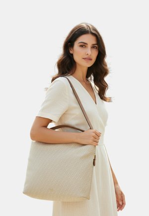 Donna con lunghi capelli scuri e mossi indossa un vestito color crema e porta sulla spalla una grande borsa tote beige con manici marroni.