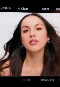 Gros plan sur une femme aux longs cheveux foncés, portant des boucles d'oreilles argentées, les lèvres légèrement entrouvertes, sur un fond clair uni.