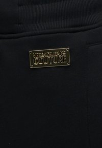 Κοντινό πλάνο μαύρου υφάσματος με μια μικρή ορθογώνια χρυσή μεταλλική πλάκα χαραγμένη με "Versace Jeans Couture", προσαρτημένη κοντά σε ριμπ υφασμένη ζώνη μέσης.