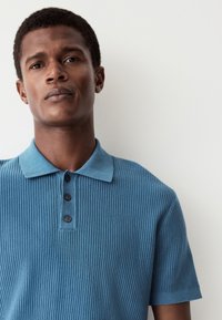 Kortärmad pikétröja i texturerat blått tyg med ribbad mönstring, utrustad med krage, tre knappar och en ren kantfinish.