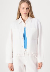 Bela denim jakna z dvema sprednjima žepoma, ovratnikom in gumbom za zapiranje. Nosi se čez modro majico, ki ima sproščen kroj in naramnice za prilagajanje.